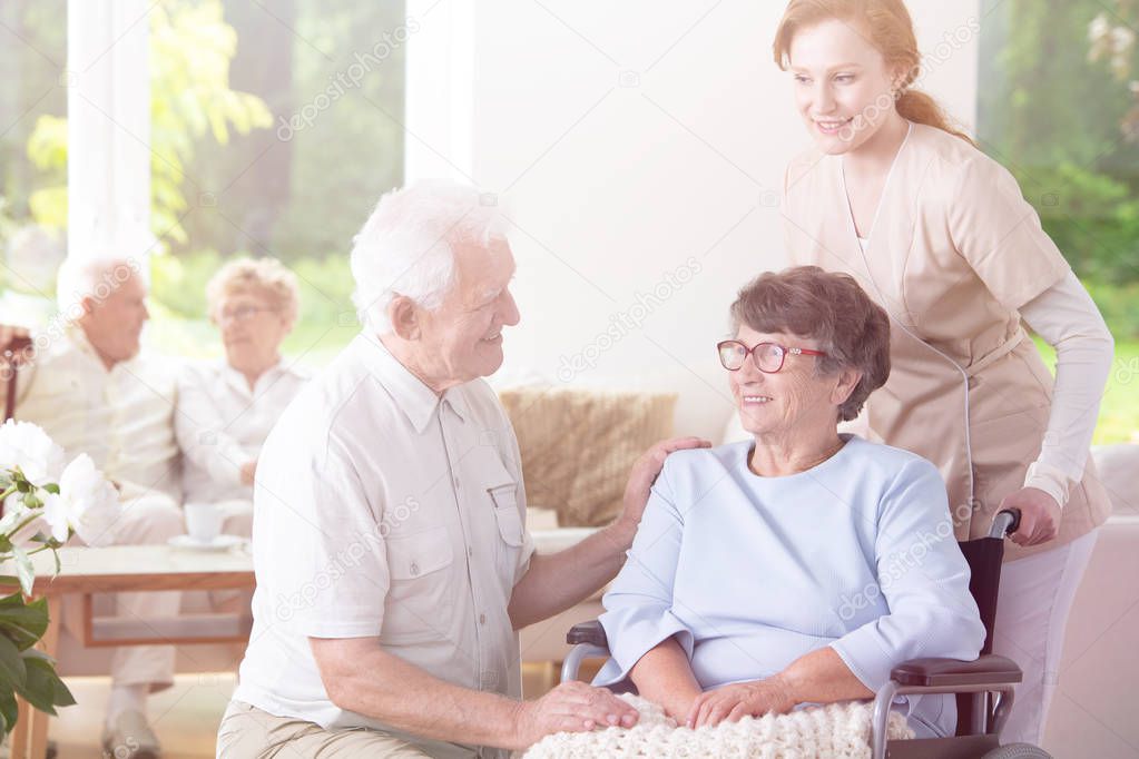 Smiling elderly man visiting happy senior woman in the wheelchair supported by nurse