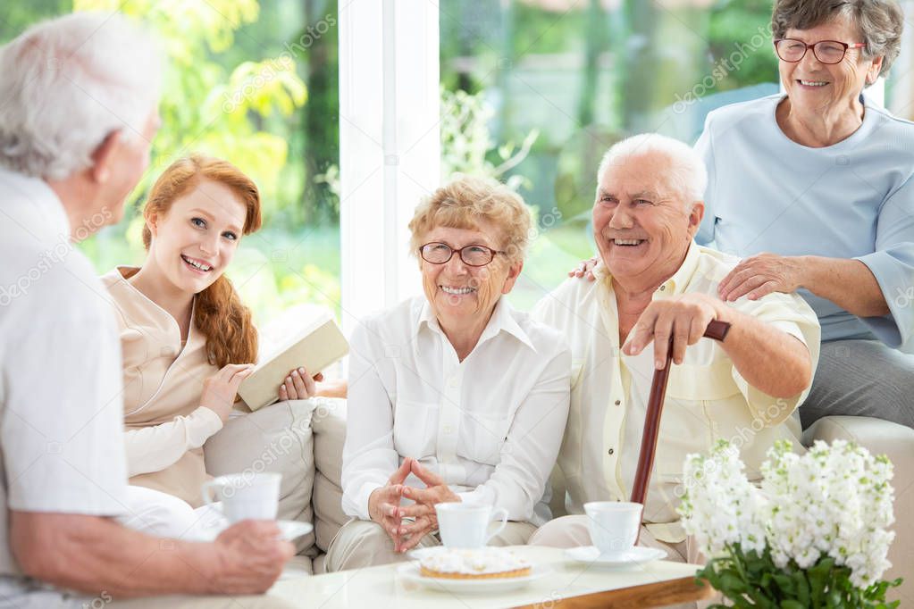 Smiling senior people drinking tea with caregiver in the common room of nursing house