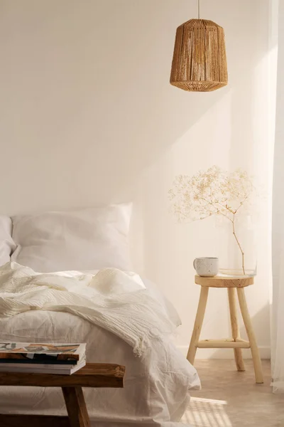 Bedside table with mug and flower next to bed with white bedding, pillow and blanket, real photo with copy space on the empty wall