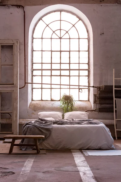 Wooden bench on carpet near bed with blanket in spacious industrial bedroom interior with window. Real photo