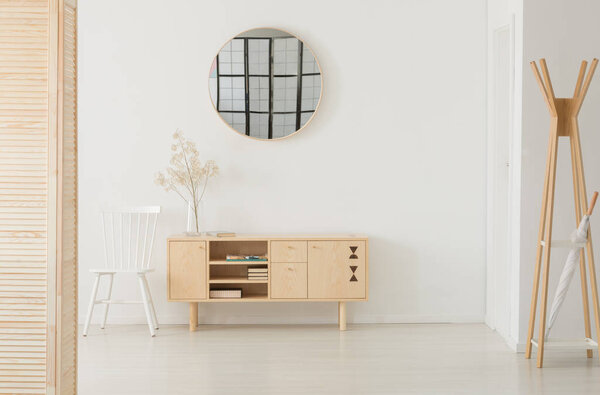 Круглое зеркало над деревянным шкафом с книгами и стеклянной вазой с цветком, рядом белый стул, настоящая фотография с копировальным пространством
