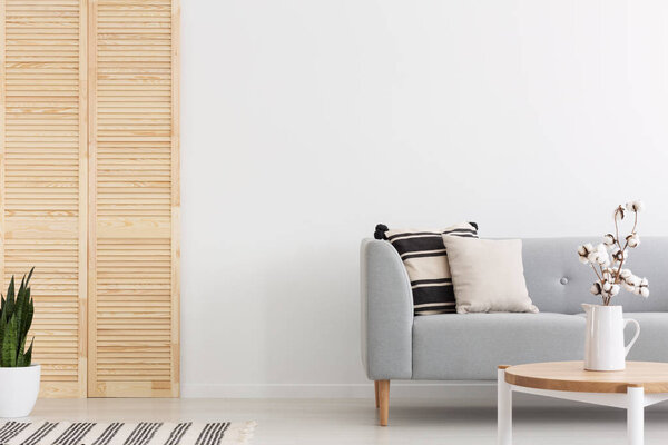 Flowers on wooden table in front of grey couch with pillows in white flat interior with plant. Real photo