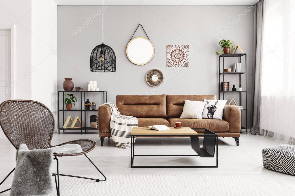 Chair near table in grey apartment interior with mirror and poster on the wall above leather sofa. Real photo