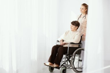 Smiling caregiver supporting paralysed elderly woman in the wheelchair next to copy space clipart