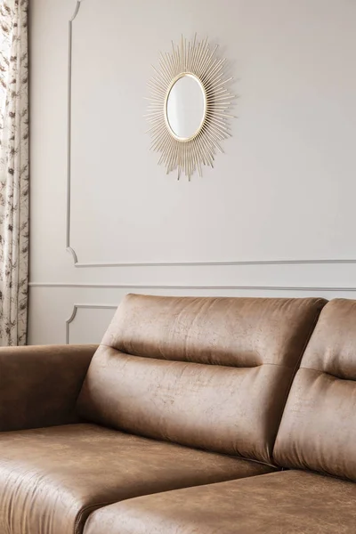 Round gold mirror hanging on the wall with molding in bright sitting room interior with leather brown couch in the real photo