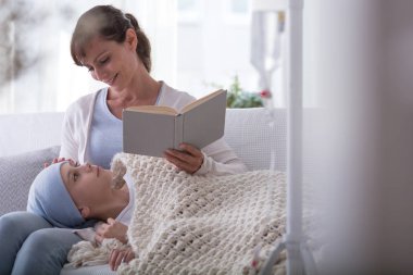 Smiling mother reading book to sick child with cancer wearing headscarf clipart