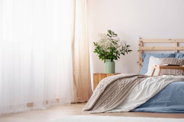 Parlak yatak odası iç mavi ve gri çarşaflı yatağın yanındaki ahşap masaya çiçek. Gerçek fotoğraf