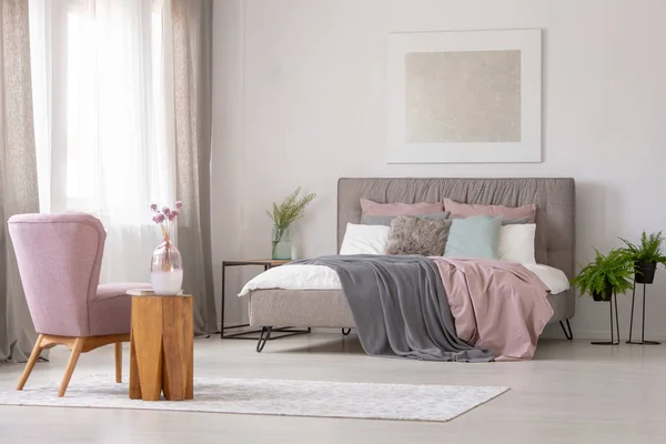 ベッドの上のポスターと灰色の寝室のインテリアにピンクのアームチェアの横に木製のテーブルの上に花 実際の写真 — ストック写真