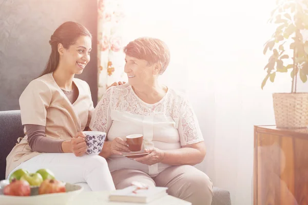 养老院的老年患者和她的护士坐在沙发上一起喝茶 — 图库照片