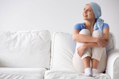 Happy teenage girl suffering from lung cancer, wearing blue headscarf and siting on the couch at hospice clipart