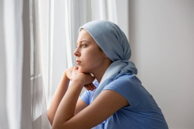 Thoughtful young girl suffering from ovarian cancer, wearing blue headscarf and looking through the window in medical center clipart