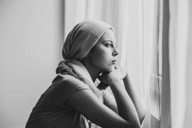 Black and white photo of thoughtful young girl suffering from ovarian cancer wearing headscarf and looking through the window in medical center clipart