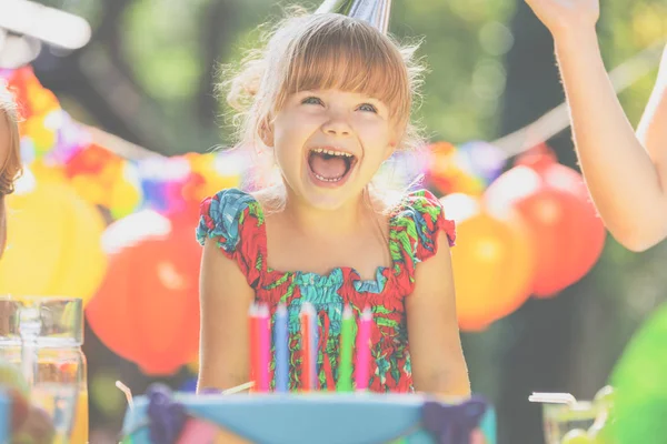 パーティーの最中に誕生日ケーキのろうそくを吹きカラフルなドレスを着て微笑んでいる女の子 — ストック写真