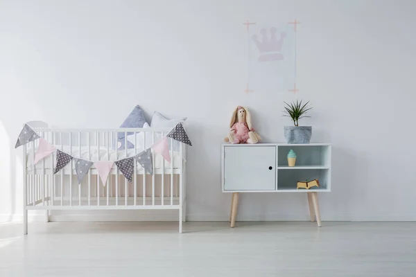 白色木制婴儿床 枕头旁边的木柜与玩具和绿色裤子在灰色 复制空间在空的白色墙壁 — 图库照片