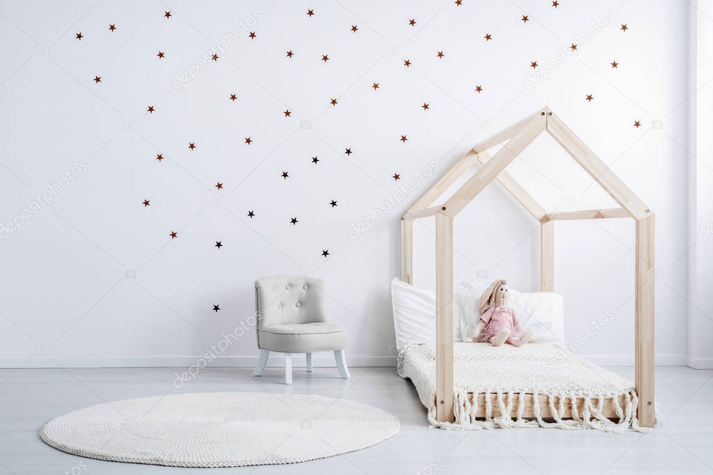 Pastel grey armchair next to wooden house shape bed with toy and blanket, copy space and golden stars on empty white wall, round white carpet on floor