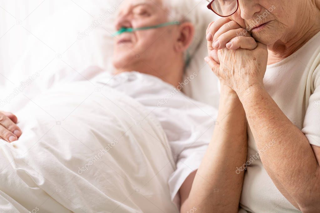Sad senior woman holding hand of her ill husband lying in hospital bed