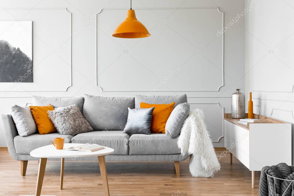 Orange lamp above table and grey settee with cushions in bright flat interior with poster. Real photo