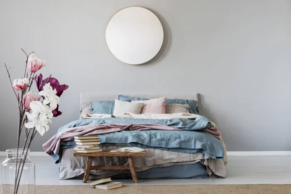 上品ラウンド ブルー パステル ピンク ベージュの寝具と暖かいベッドでスタイリッシュな寝室のインテリアに灰色の壁に鏡と木製のベンチの本 — ストック写真