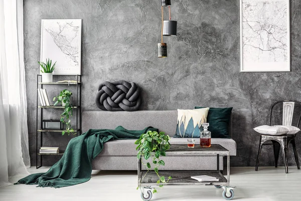 Dark green blanket on comfortable grey sofa in elegant industrial living room with maps on empty wall