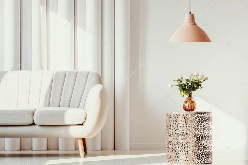 Pastel orange lamp above white flowers in glass vase on trendy openwork table in bright living room interior with white couch