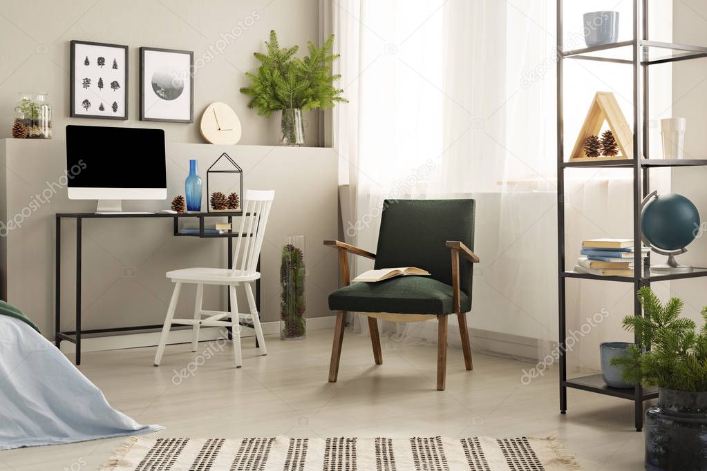 Open book on dark green retro armchair in the middle of stylish workspace with metal shelf with books and desk with all in one computer