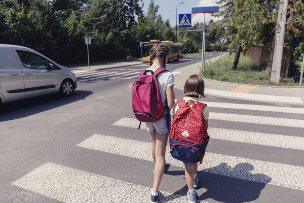 Children with backpacks walking to the school through a crosswalk next to a car
