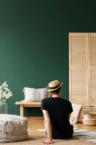 穿着黑色 T恤和柳条帽的希普斯特坐在天然客厅的地板上 配有木制家具 真正的照片 空绿色墙上有复制空间 — 图库照片