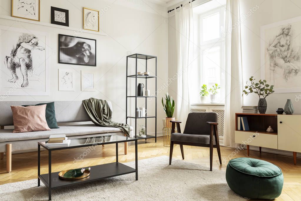 Pouf next to armchair in living room interior with table in front of sofa under posters. Real photo