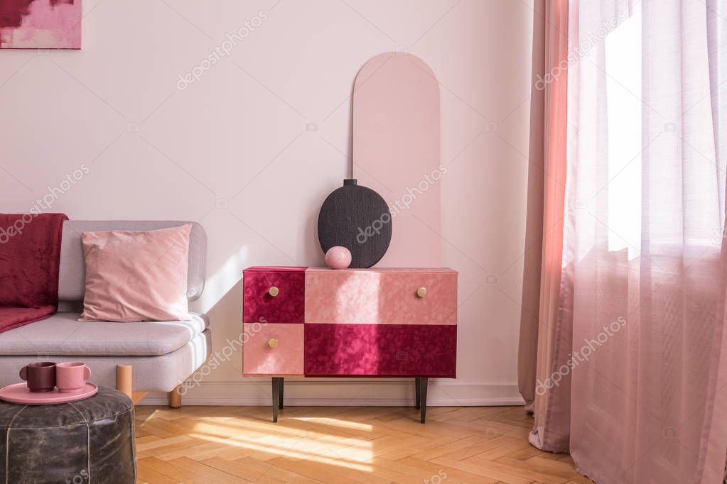 Elegant suede covered pastel pink and burgundy colored cabinet with golden handles next to window in bright living room interior