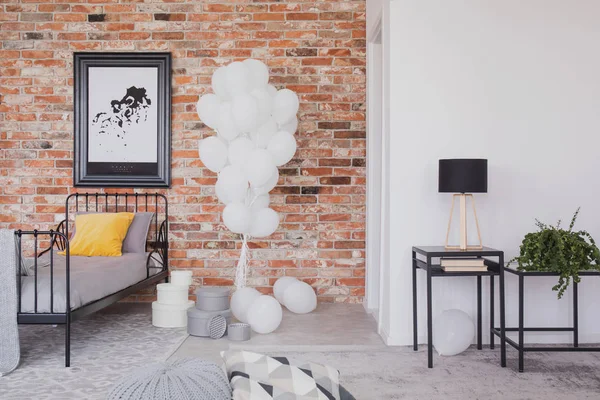 Bunch of white balloons next to single black metal bed with grey bedding and yellow pillow in industrial bedroom interior with brick wall
