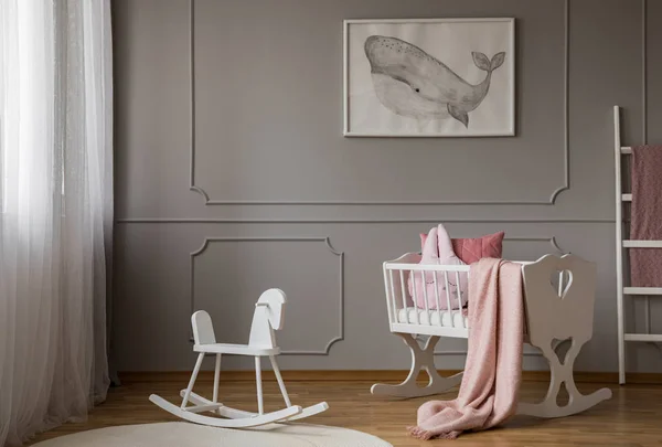 Rocking horse on rug next to cradle in grey kid\'s bedroom interior with whale poster. Real photo