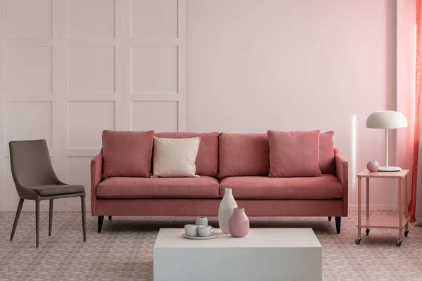 Stylish living room interior with pastel pink velvet sofa with pillows and trendy grey chair