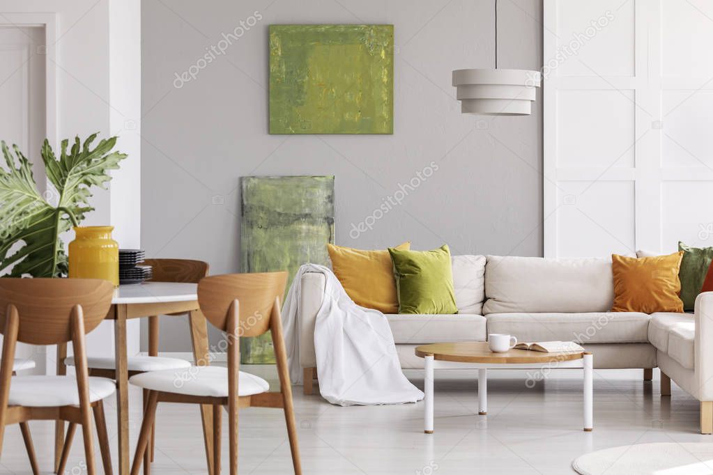 Wooden chairs at table in grey flat interior with pillows on corner couch near green posters. Real photo