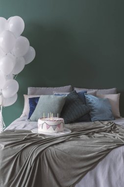 Doğum günü pastası ile rahat çift kişilik yatak yastık ve battaniye, kopya alanı boş yeşil duvar ve beyaz balonlar köşedeki demet ile mum