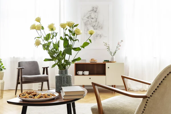 Armchair at table with flowers, books and food in bright apartment interior with windows. Real photo