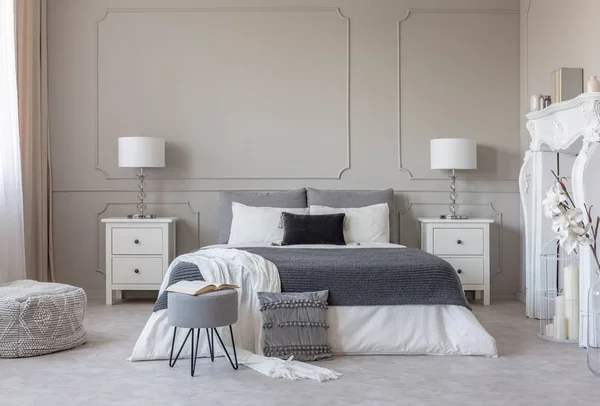 Grey pouf with open book in the food of king size bed with grey and white bedding, copy space on empty wall