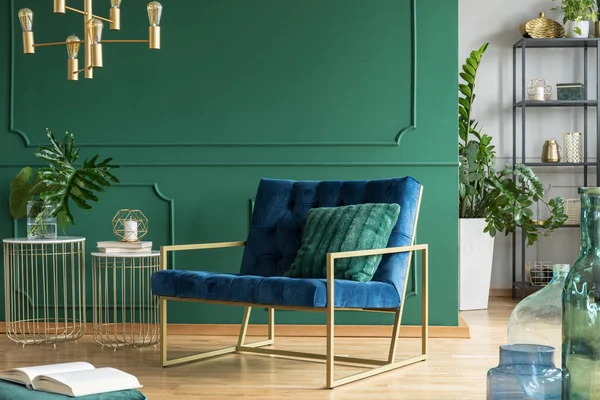 Petrol blue armchair with green pillow next to two stylish coffee tables, copy space on empty green wall