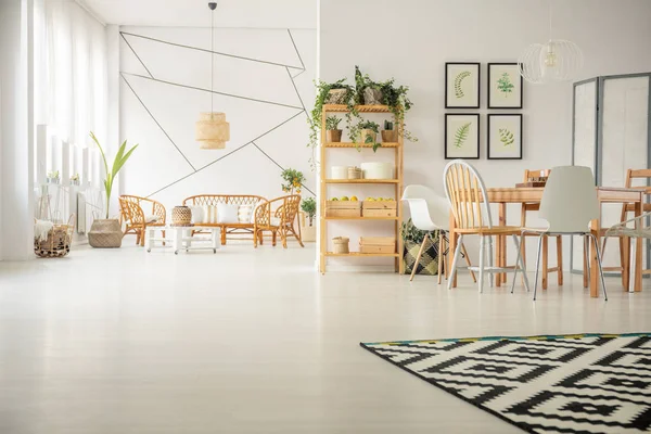 Patterned carpet on white wooden floor in elegant living and dining room interior with rattan sofa and armchairs and wooden table with fashionable chairs