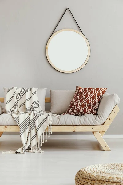 Round mirror in wooden frame on the wall of bright beige living room with comfortable sofa with patterned pillow and striped blanket, real photo