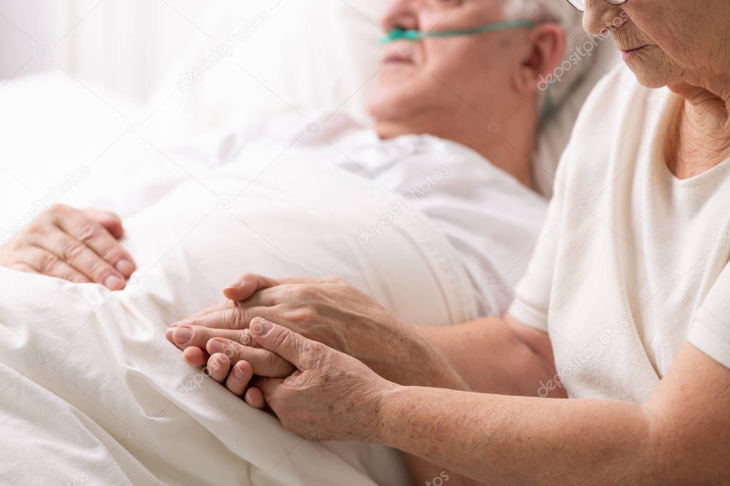 Senior grandmother holding hand of her cancer sick husband lying in white hospice bed, photo with copy space