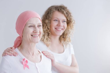 Smiling senior woman suffering from tumor wearing pink headscarf and ribbon, her daughter standing behind her clipart