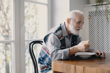 Gri saçlı ve sakallı yaşlı Büyükbaba mutfakta yalnız oturuyor kahvaltı yiyor