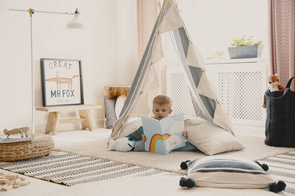 Cute child reading a book, laying on the pillows in stylish scandinavian tent in playroom