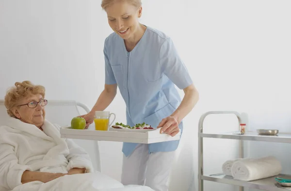 Happy carer in blue uniform with tray with glass of orange juice and portion of fruits and senior woman lying in hospital bed