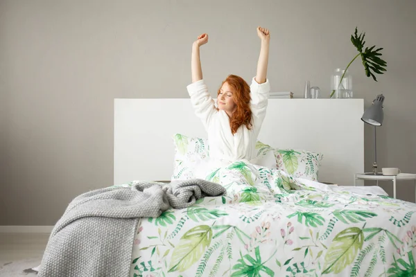 花柄の寝具と灰色の毛布 灰色の寝室の設計の実際の写真で大きな快適なベッドで目を覚ます笑顔赤毛の女の子 — ストック写真