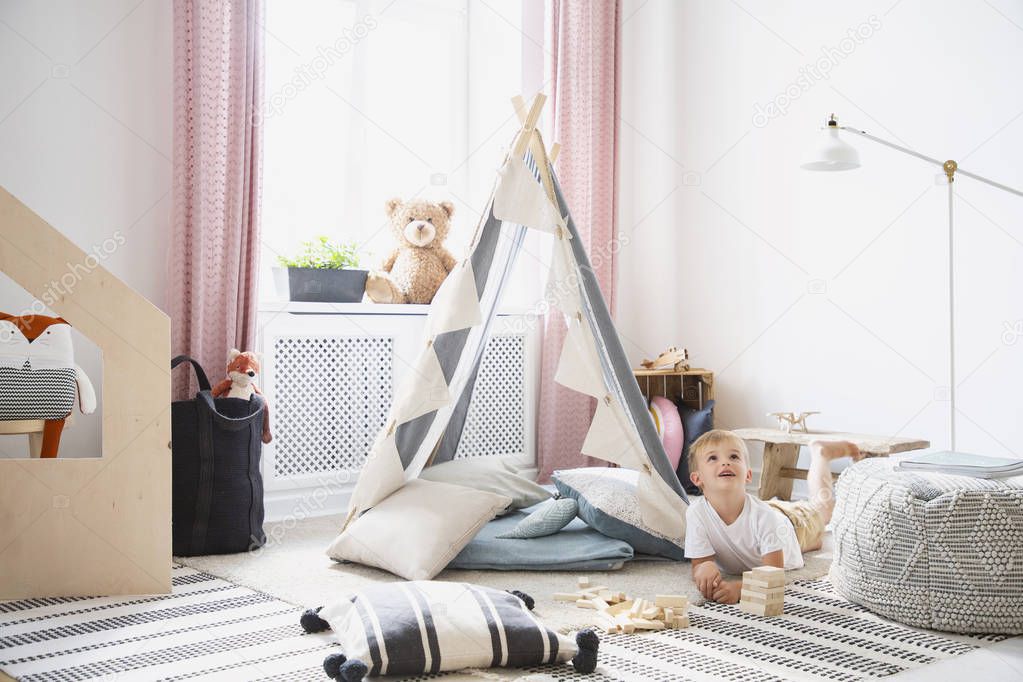 Happy child playing on the floor between pouf and tent in bright playroom interior with lamp. Real photo