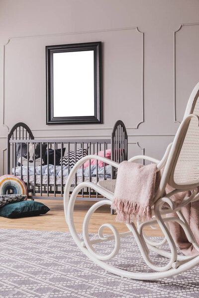 White rocking chair with pastel pink blanket in elegant little baby bedroom with grey wooden crib and mockup poster on the wall