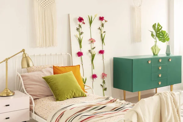 Цветочная доска между односпальной кроватью с оливково-зелеными, оранжевыми и розовыми подушками и зеленым деревянным шкафом с листочком в стеклянной вазе — стоковое фото