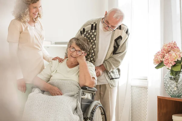 Парализованная жена и ее муж улыбаются в доме престарелых при содействии медсестры — стоковое фото