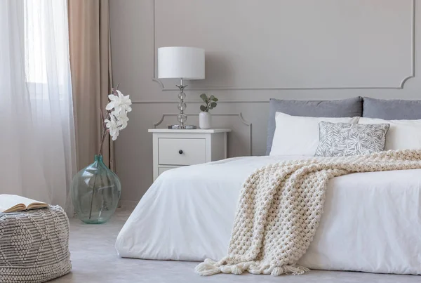 Witte stijlvolle lamp op houten nightstand naast bloem in grote glazen vaas in eigentijdse slaapkamer interieur — Stockfoto